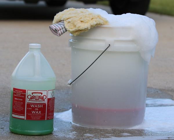 A white bucket overflowing with soap suds with a wash mitt on top. A gallon jug of Showcar Wash-N-Wax beside it. Driveway background